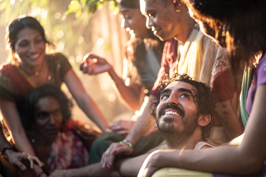 A film still of Dev Patel, smiling up at a woman. He is lying in her lap, with other people seated around them.