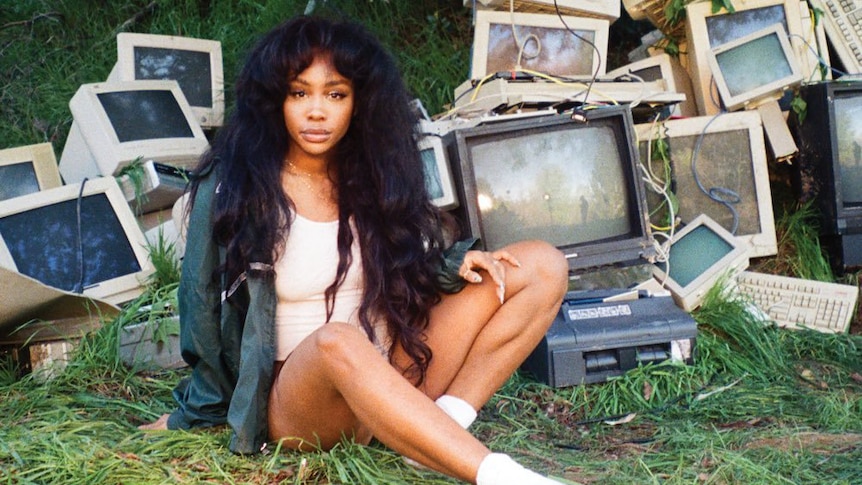 SZA Sits on grass in front of a pile of old computers and CRT Monitors