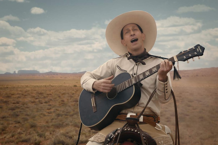 Man on horseback wearing light-coloured cowboy hat plays guitar and sings, Western plains behind him and blue sky above.