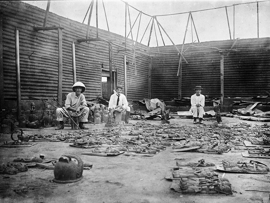 Three men sitting in the ruins of a large room. On the floor are dozens of bronze plaques and sculptures.