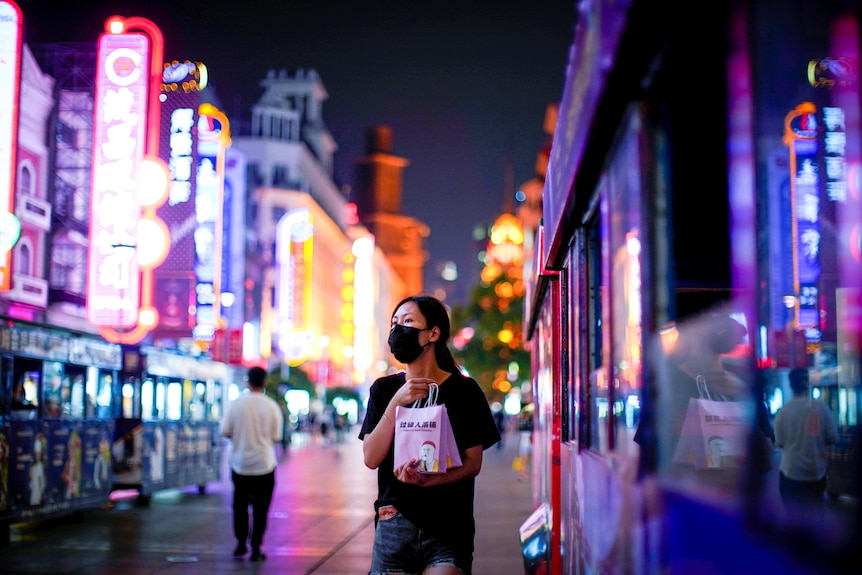 A young chinese woman in a face mask on a street bathed in fluorescent lights in Shanghai