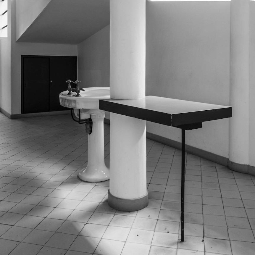 A black and white photo shows a sink in the middle of a modernist hall with tiled floors and sweeping white columns.