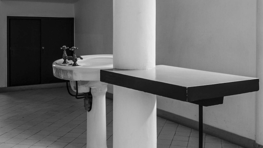 A black and white photo shows a sink in the middle of a modernist hall with tiled floors and sweeping white columns.