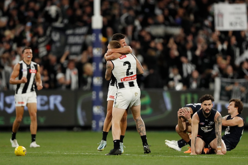 Two Collingwood AFL players embrace in the middle of the field as a trio of Carlton players sit dejected on the ground. 