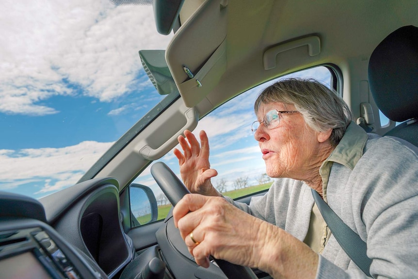 An older woman in the driver's seat of a car gestures towards the windshield with one hand, her other hand steering the wheel.