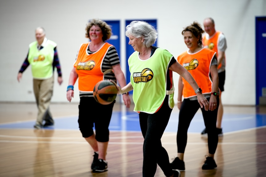 Woman with grey hair and basketball, smiling and bouncing a basketball on a court. She's playing walking basketball with others.