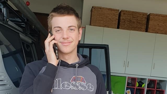 A young man with short brown hair and a moustache, wearing a hooded jumper, smiles while on the phone.