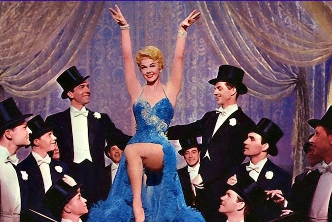 Doris Day wears a blue evening gown surrounded by men in black suits and top hats.