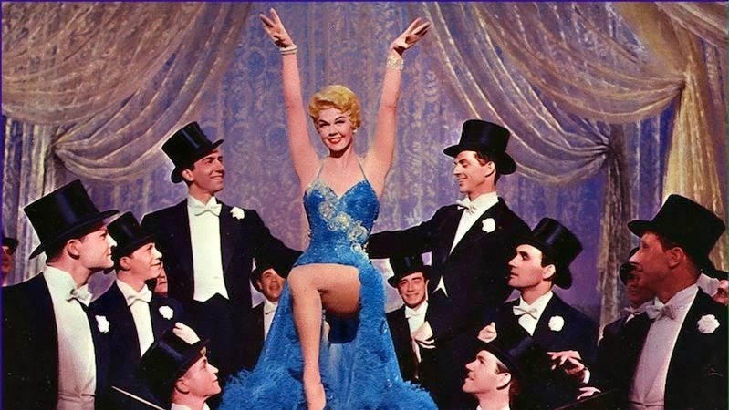 Doris Day wears a blue evening gown surrounded by men in black suits and top hats.