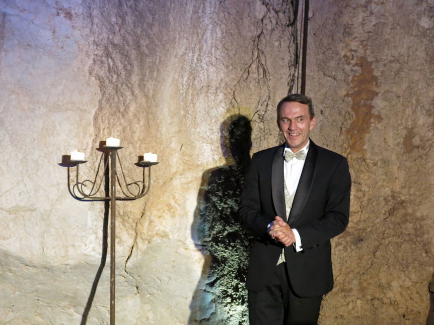 A man in a tuxedo stands next to a stand of candles, with limestone rock behind him