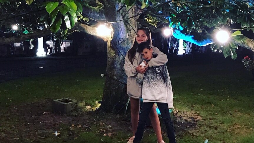 A sister hugs her little brother under while standing under a tree festooned with fairy lights.