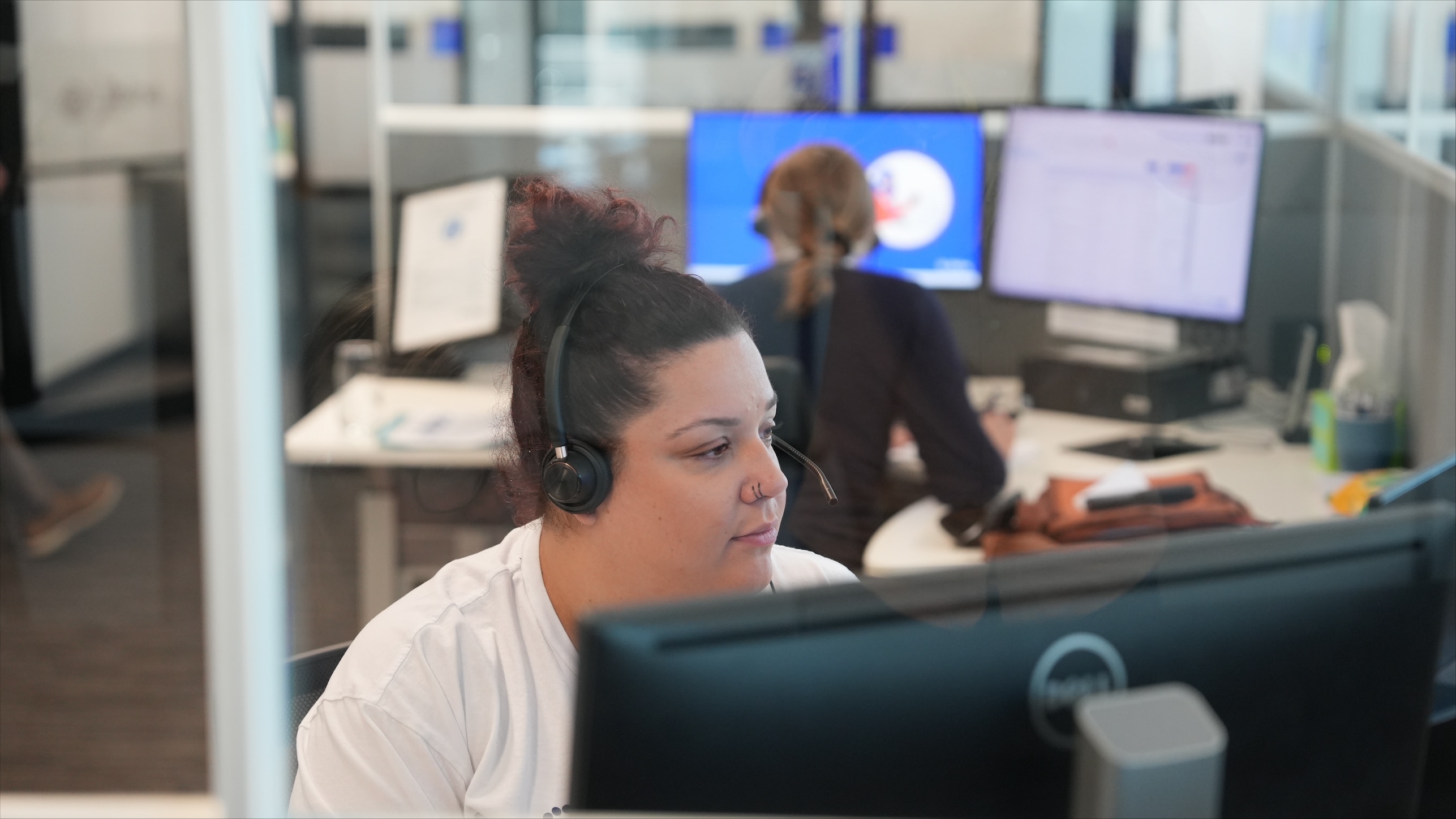 A person with dark hair sitting in glass cubical at a computer, wearing a pair of headphones with a built-in microphone.