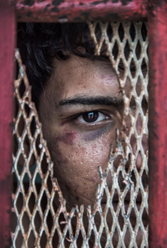 Behind a worn maroon metal door with broken wire grate peers a man with facial marks and bruises, only half of his face is seen.