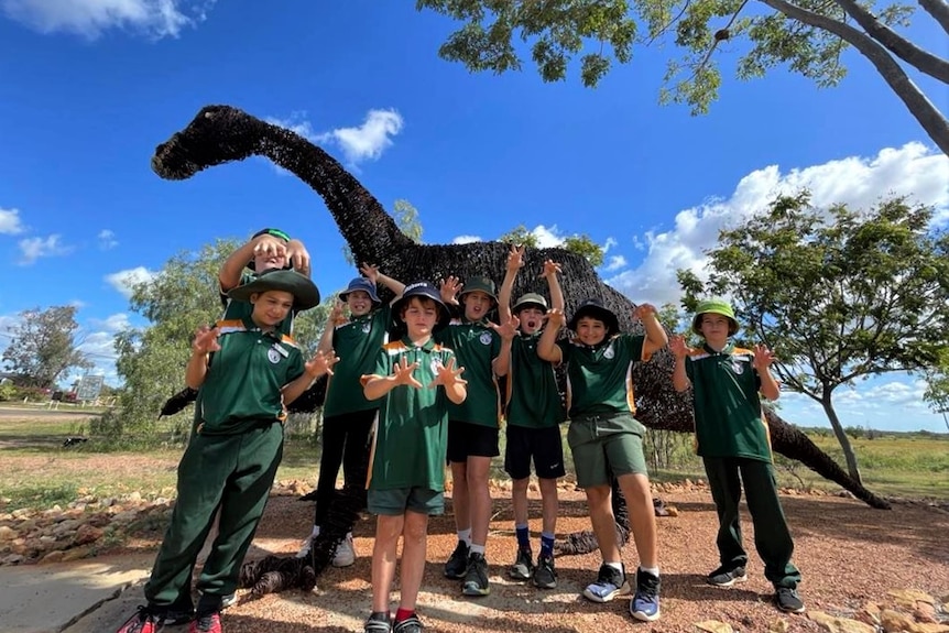 Primary school kids in green uniforms stand on red soil in front of an enormous dinosaur statue in their town