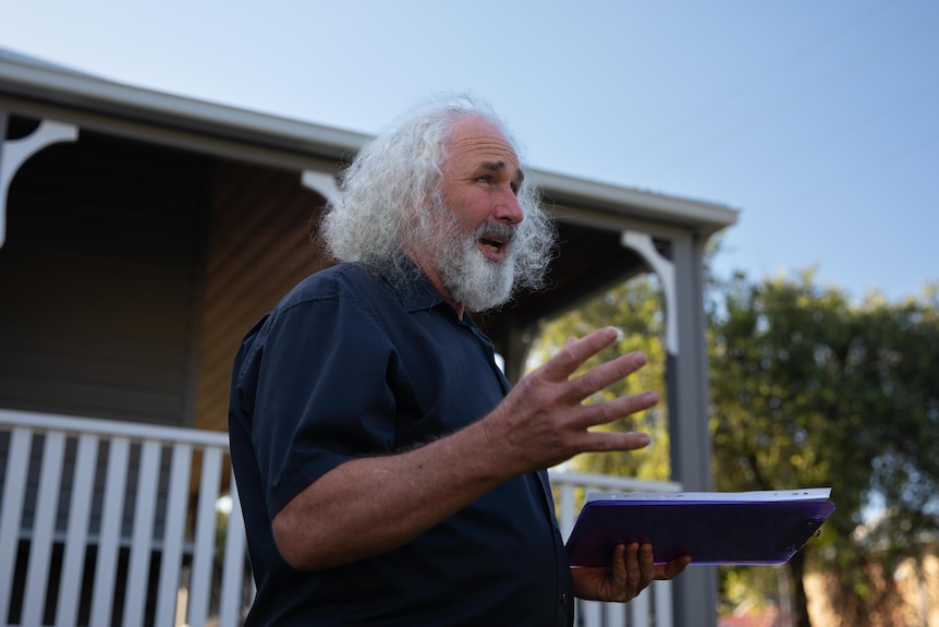 Man with should-length white hair and beard gestures with his hand 