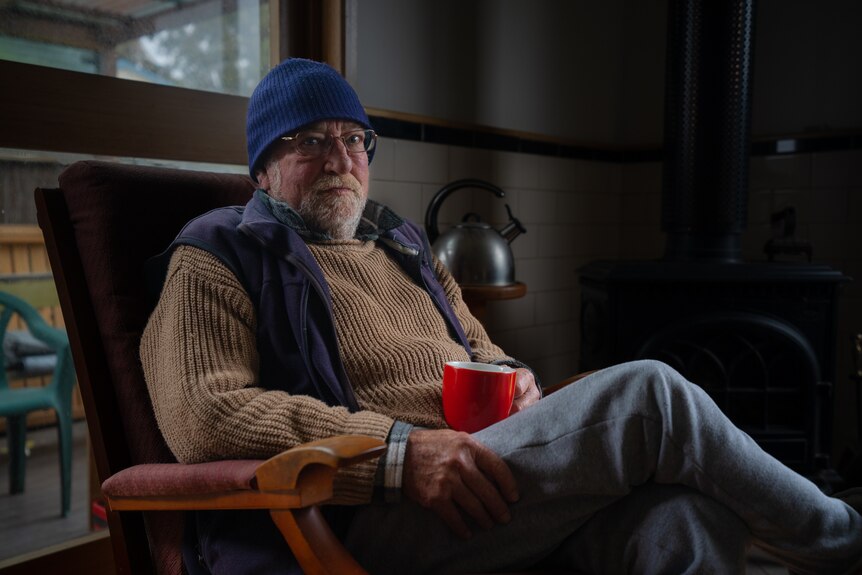 A man in a beanie holds a cup of tea and looks at the camera.