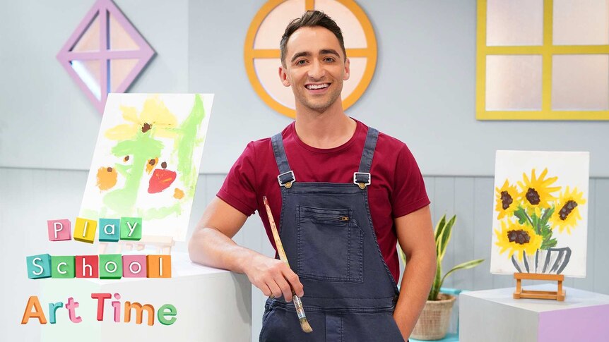 Art Time host is holding a brush in one hand and has the other in one pocket of his blue apron