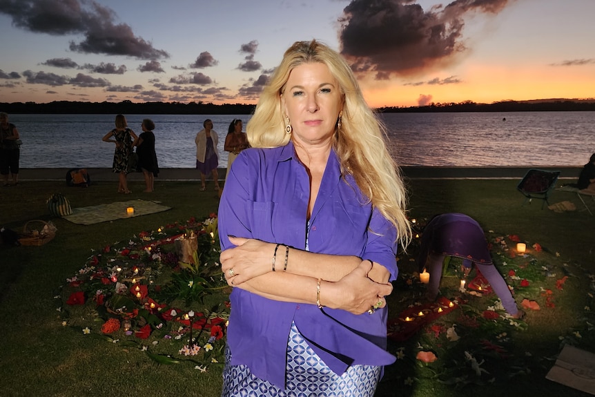 A woman with a purple shirt stands in front of a floral display near a river shortly after sunset.