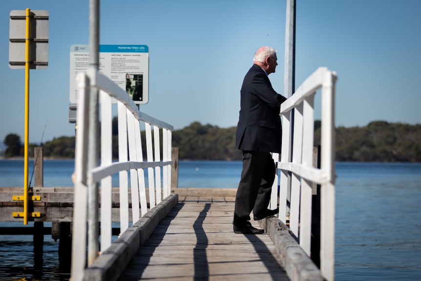 An older man stands on a jetty looking out at the river. 