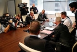 A group of people sit around a table with documents and computers in front of them while camera operators capture proceedings.