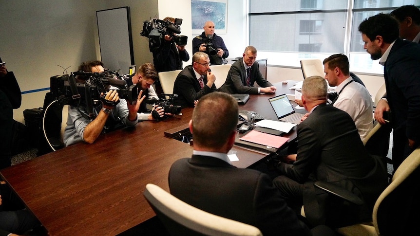 A group of people sit around a table with documents and computers in front of them while camera operators capture proceedings.