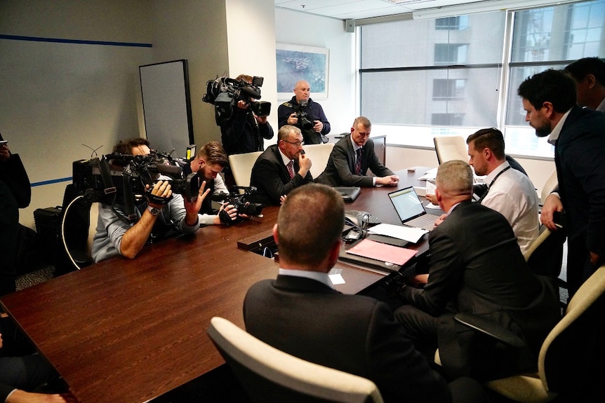 A group of people sit around a table with documents and computers in front of them while camera operators capture proceedings.