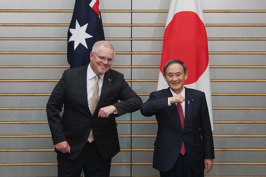 Two men smile while bumping elbows in greeting in front of an Australian and Japanese flag