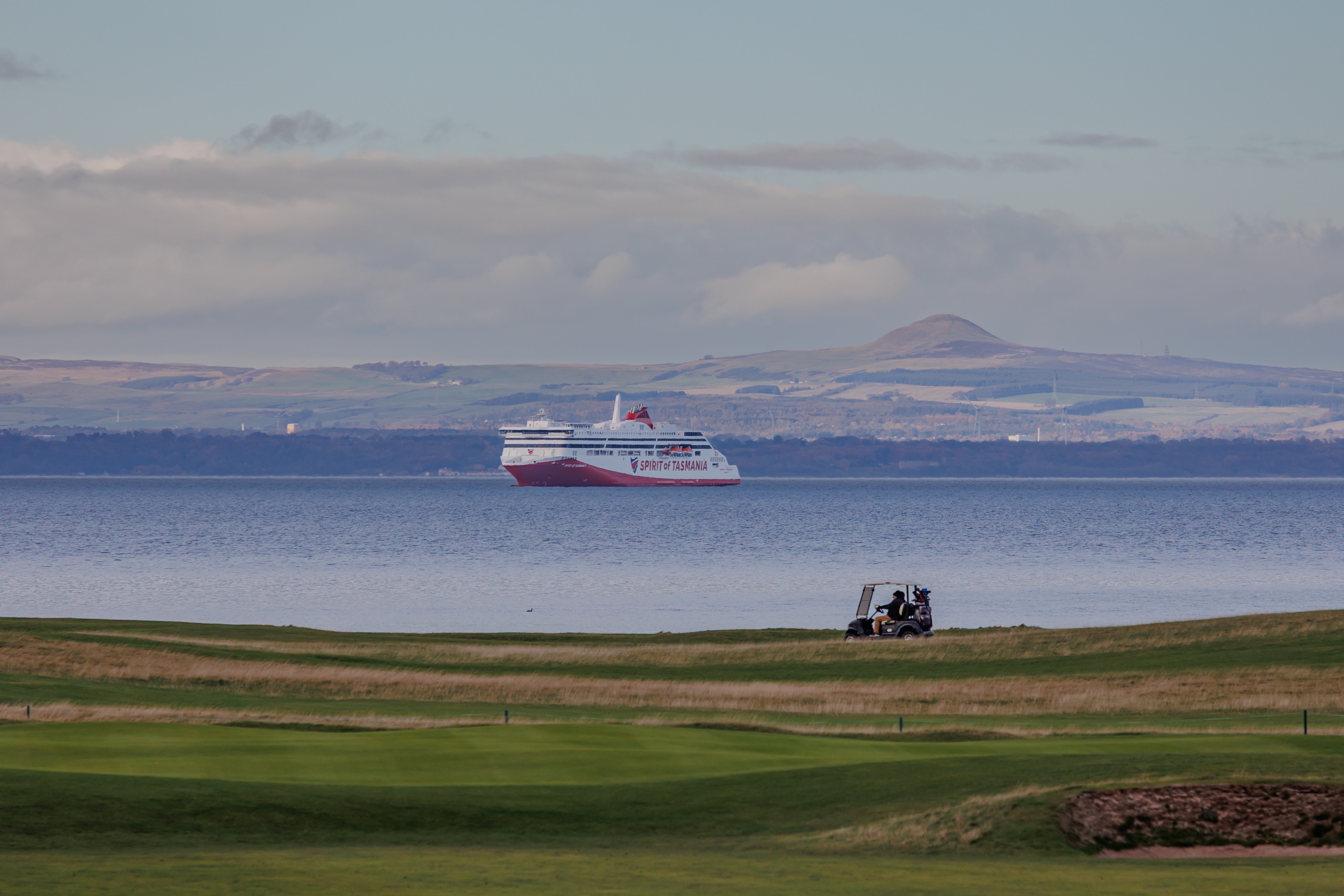 A large red and white ferry sails past a golf course.