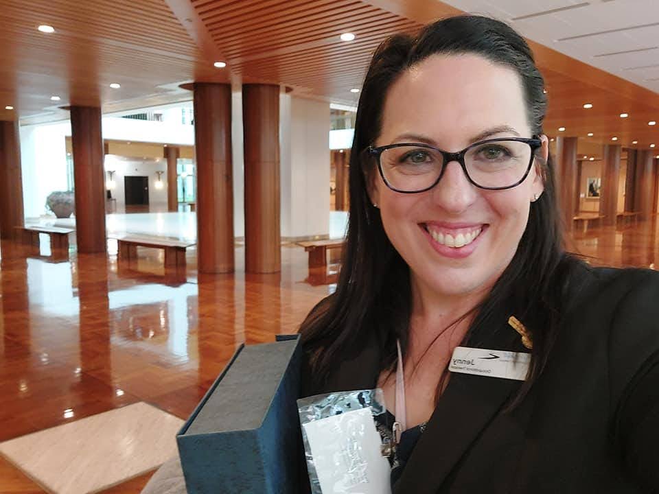 A woman with dark brown hair and black framed spectacles takes a selfie in a formal foyer with timber ceiling and polished floor