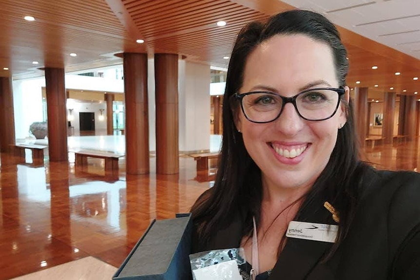 A woman with dark brown hair and black framed spectacles takes a selfie in a formal foyer with timber ceiling and polished floor