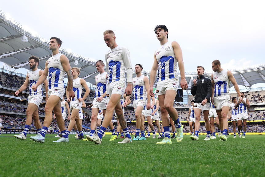 A group of dejected North Melboiurne AFL players walk off the ground after losing a game.