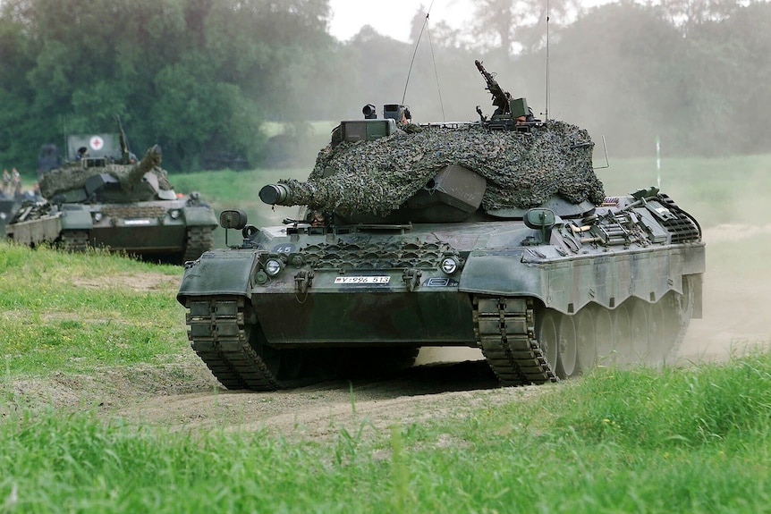 A military tank driving through a field, with another tank not far behind it.