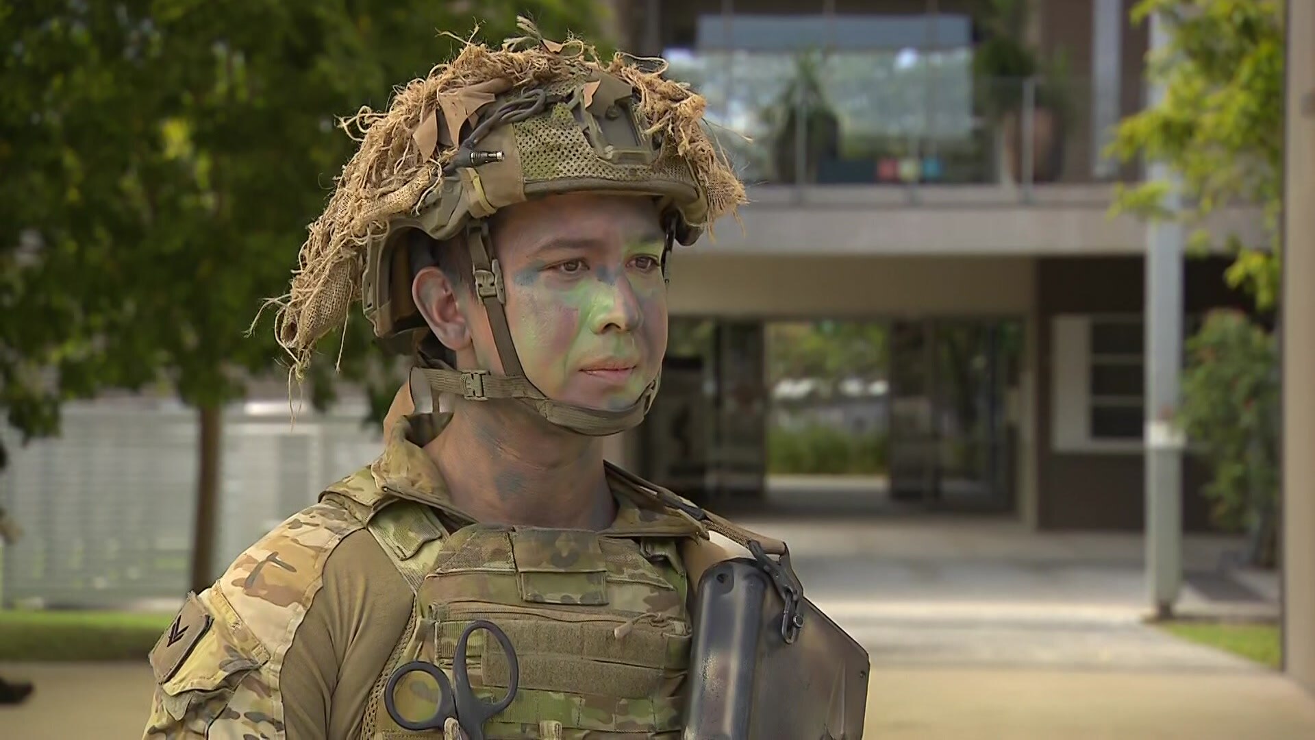 Uma fotografia da cabeça e dos ombros de um jovem soldado em um traje camuflado e com o rosto escurecido.
