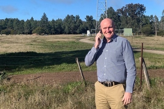 A man stands ina paddock with a phone to his ear. There is a telecommunications tower behind him.