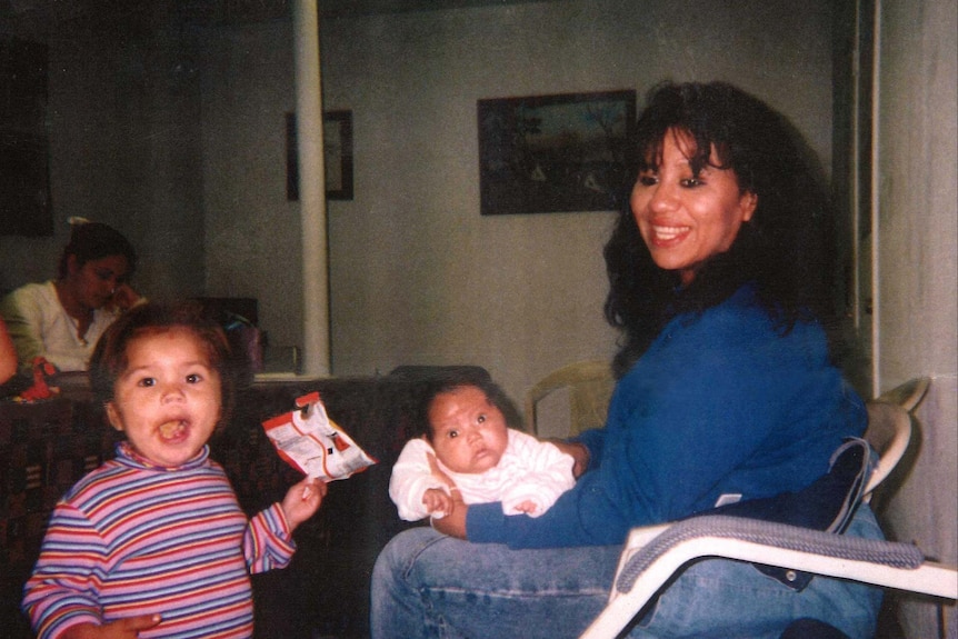 A young woman sits with on a chair with a baby on her lap, gazing at a toddler standing next to her smiling at camera
