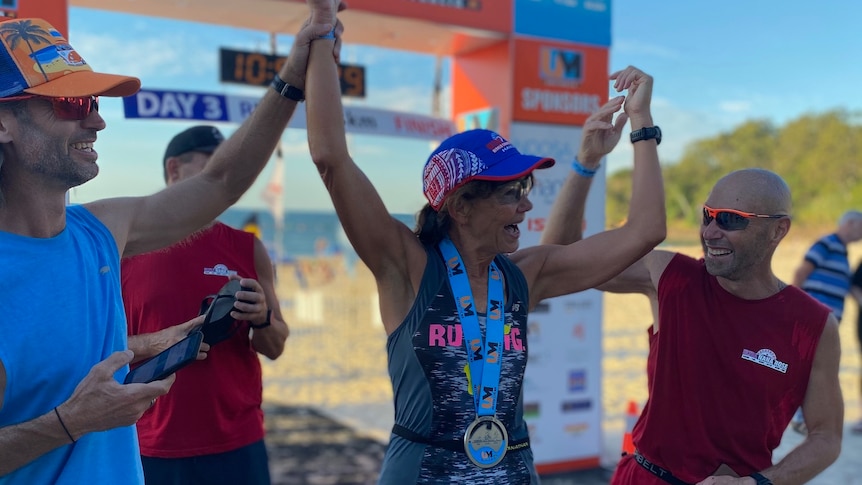 A very fit woman wearing a cap and a medal around her neck at the finished line, raises her arms, flanked by two supporters.