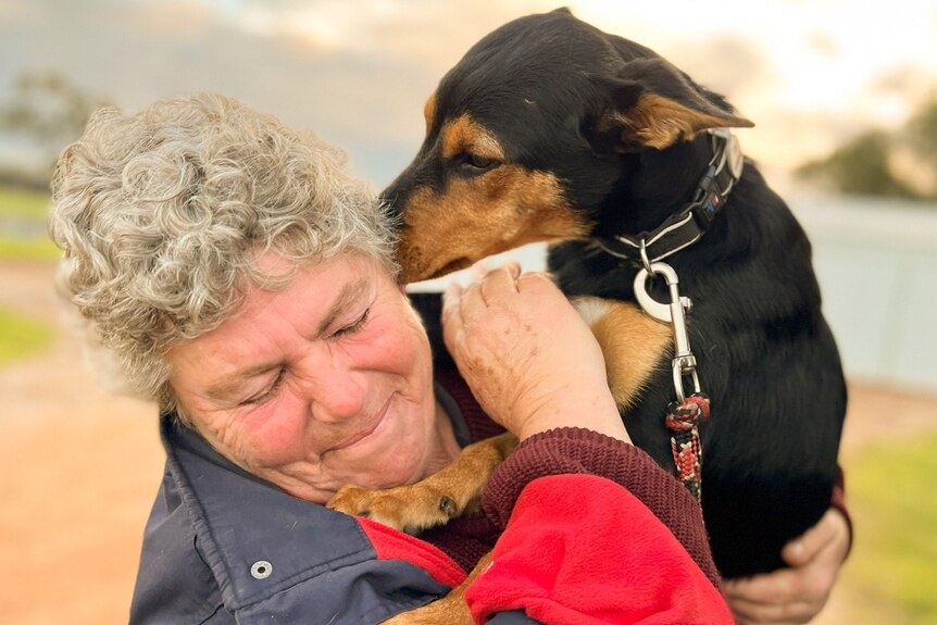A woman with curly grey hair smiles as she holds a brown and black dog to her face, cuddling.