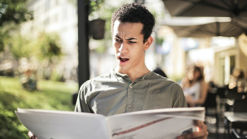 A man who is reading a newspaper has a shocked expression on his face