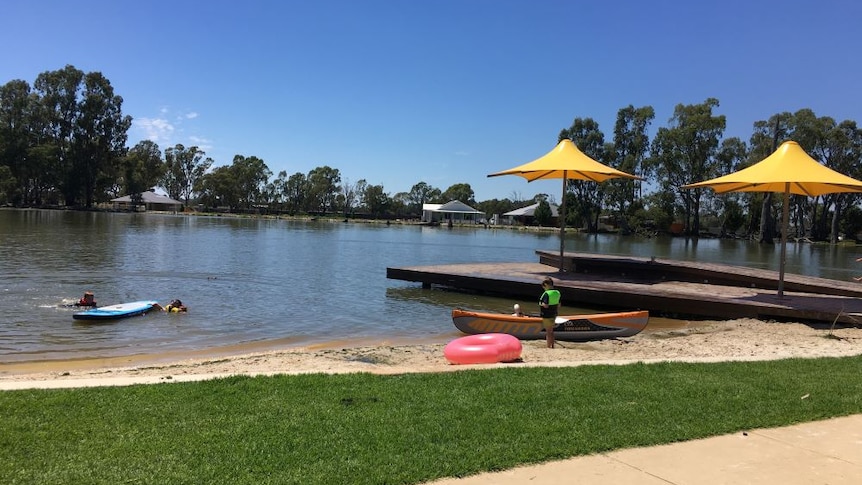 A boy stands beside a kayak on the sand, while two kids are in the Gunbower Creek next to a stand up paddleboard.