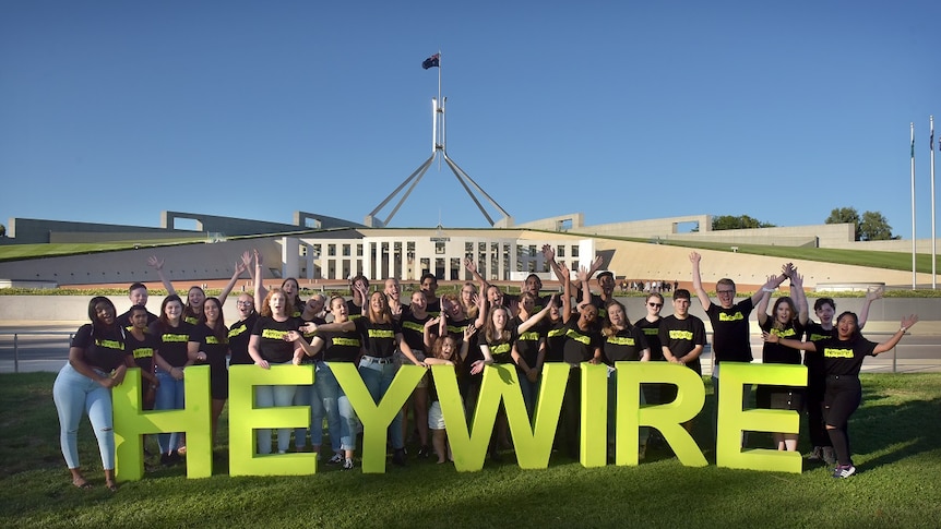 A large group of young people stand in front of Parliament House in Canberra with big green letters than spell HEYWIRE