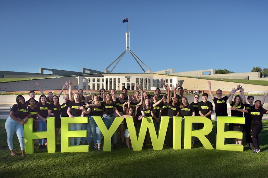 A large group of young people stand in front of Parliament House in Canberra with big green letters than spell HEYWIRE