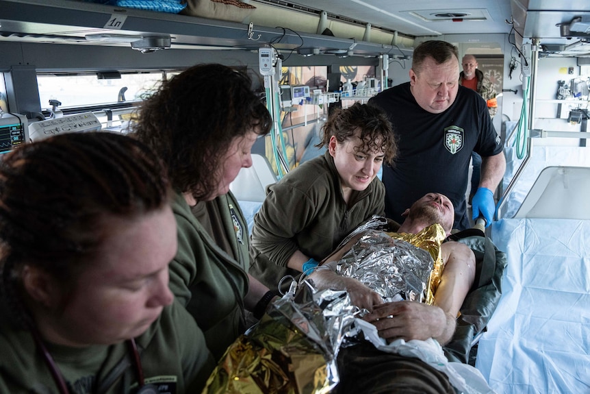 Four people carry a man on a stretcher who is covered in a silver blanket.