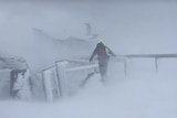 A rescue worker wearing a high vis jacket walks through a blizzard. A building is faintly visible in the distance.