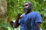Ishmael Toroama, in a blue T-shirt, stands and gestures as several men sitting nearby watch on