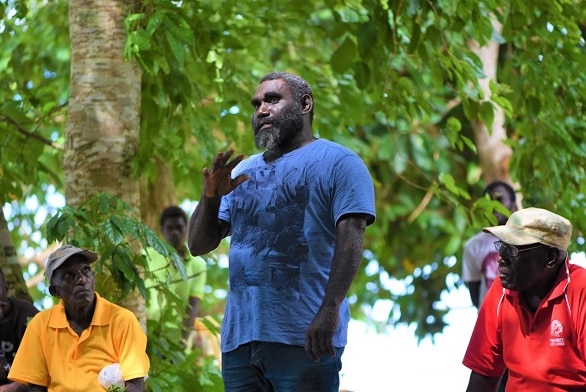 Ishmael Toroama, in a blue T-shirt, stands and gestures as several men sitting nearby watch on