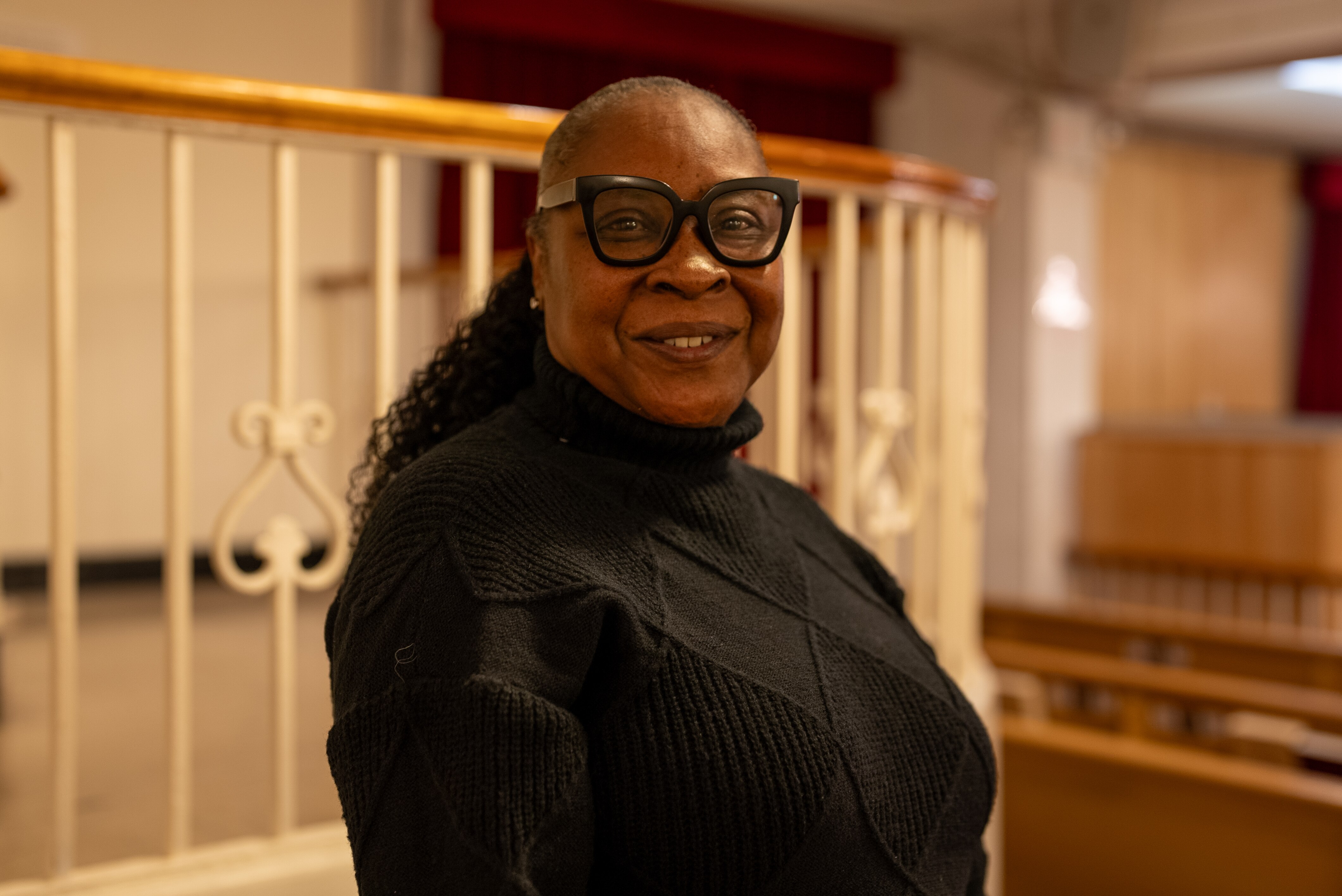 A black woman smiling in church wearing glasses and a black knitted sweater.