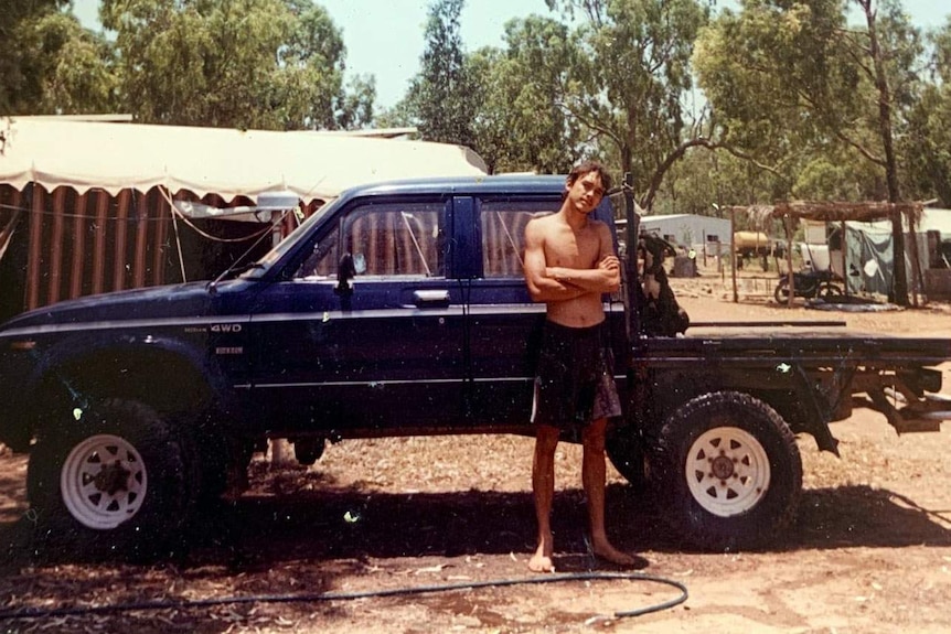A colour photograph of a young man standing in front of a trayback vehicle with his arms folded