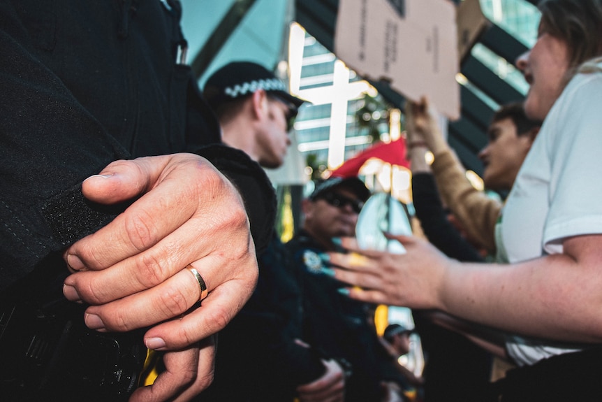 A wedding ring on a police officer's hand as he stands in front of a group of people chanting.