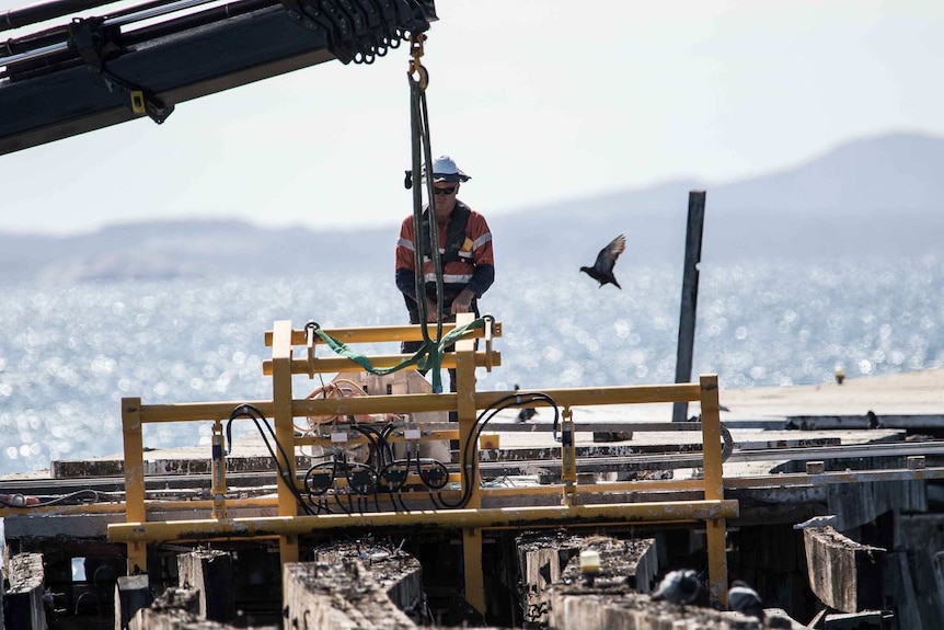 Machinery in the foreground; a jetty, worker and a glittering ocean in the background.