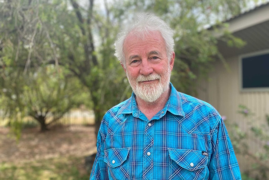 A man with white hair wears a blue flannel shirt in front of a house and garden